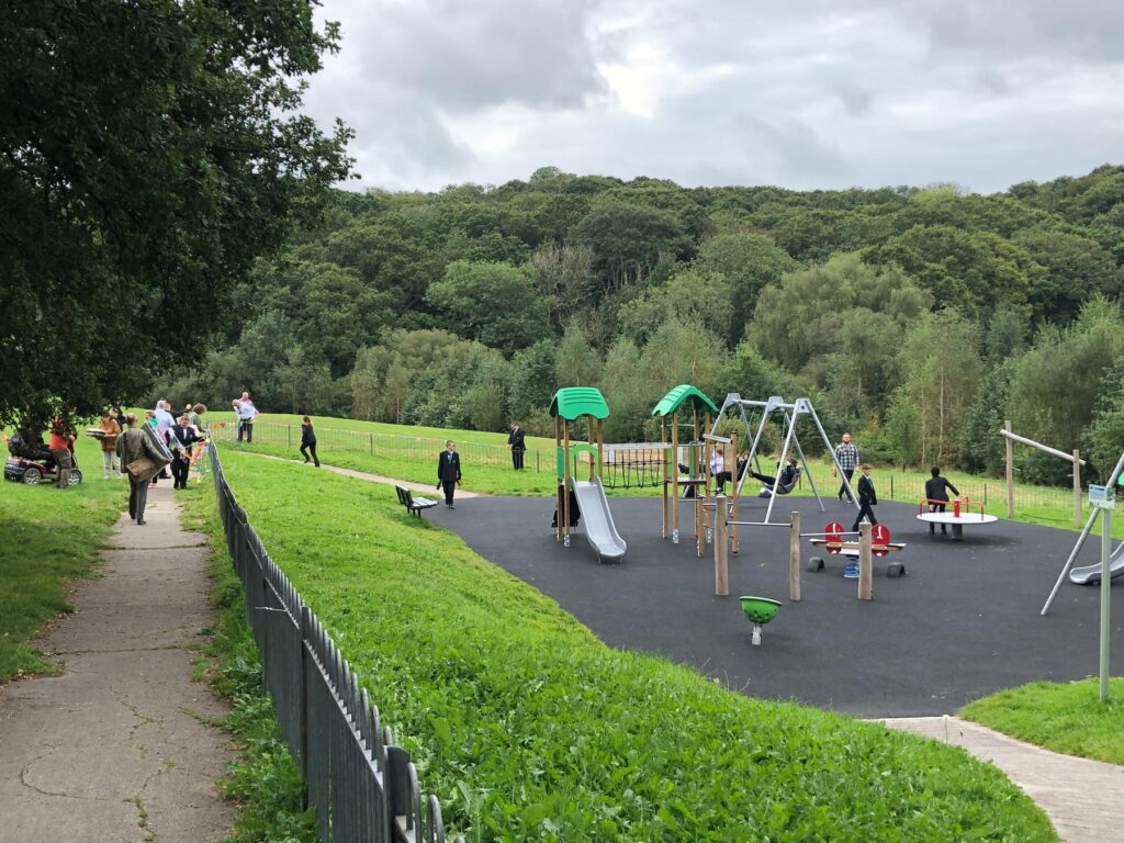 A play park with a slide situated in a grassy park surrounded by trees.