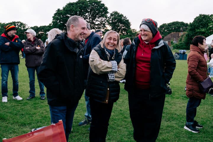 A group of three people laughing together. They are wearing dark coloured jackets and standing in a park at a community event, with some more people in the background.