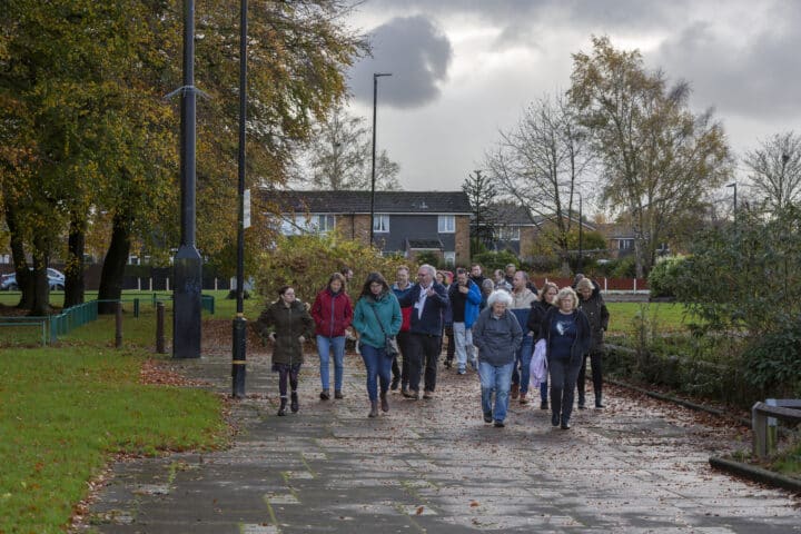 A group of people walk along a paved path in a park with grass and trees on either side. There is a grey sky and some houses in the background.