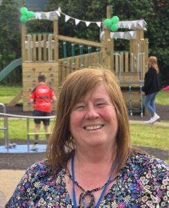 Christine is standing outdoors in front of a wooden playground decorated with green balloons and white bunting. She is wearing a floral blouse and a blue lanyard. In the background, a child in a red sports jersey is playing, and a woman in a black jacket and jeans is walking nearby.