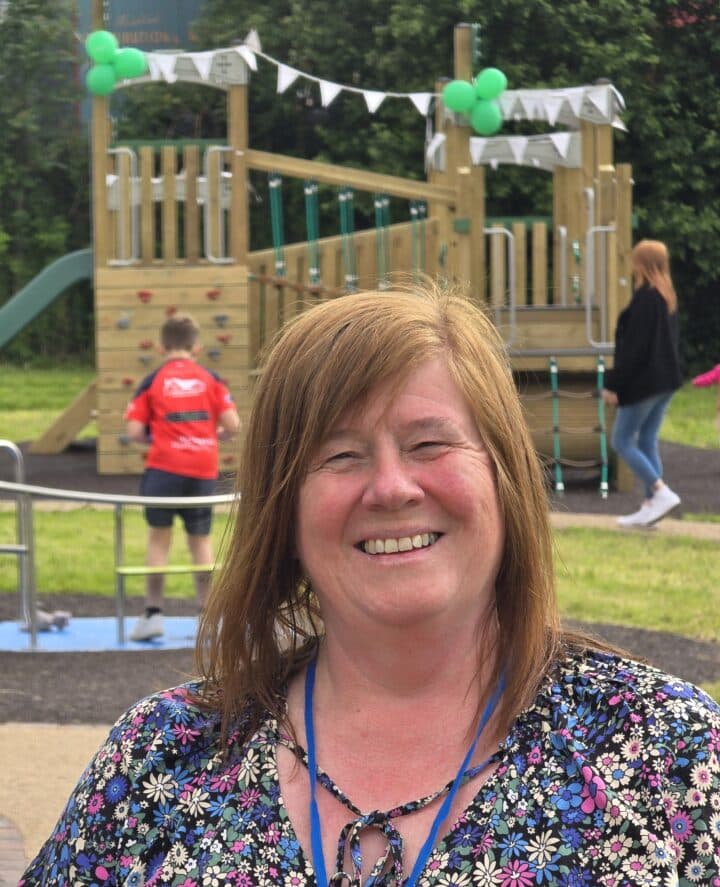 Christine is standing outdoors in front of a wooden playground decorated with green balloons and white bunting. She is wearing a floral blouse and a blue lanyard. In the background, a child in a red sports jersey is playing, and a woman in a black jacket and jeans is walking nearby.