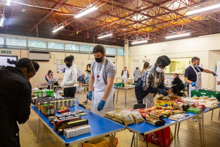 A group of volunteers wearing masks and gloves are working in a community food bank inside a hall with a high ceiling and exposed red beams. They are organiszing and distributing food items such as canned goods, pasta, and bread on blue tables. Some volunteers are wearing aprons with logos. Others in the background are packing supplies and assisting people.