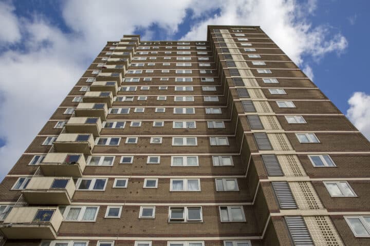 A tall brown brick high-rise apartment building with numerous white-framed windows and balconies on the left side. The photo is taken from a low angle, looking up toward a partly cloudy blue sky.
