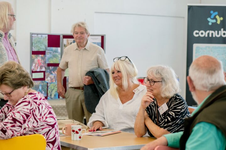 A group of older adults smiling and chatting in a community hall. Some are seated and some are standing.