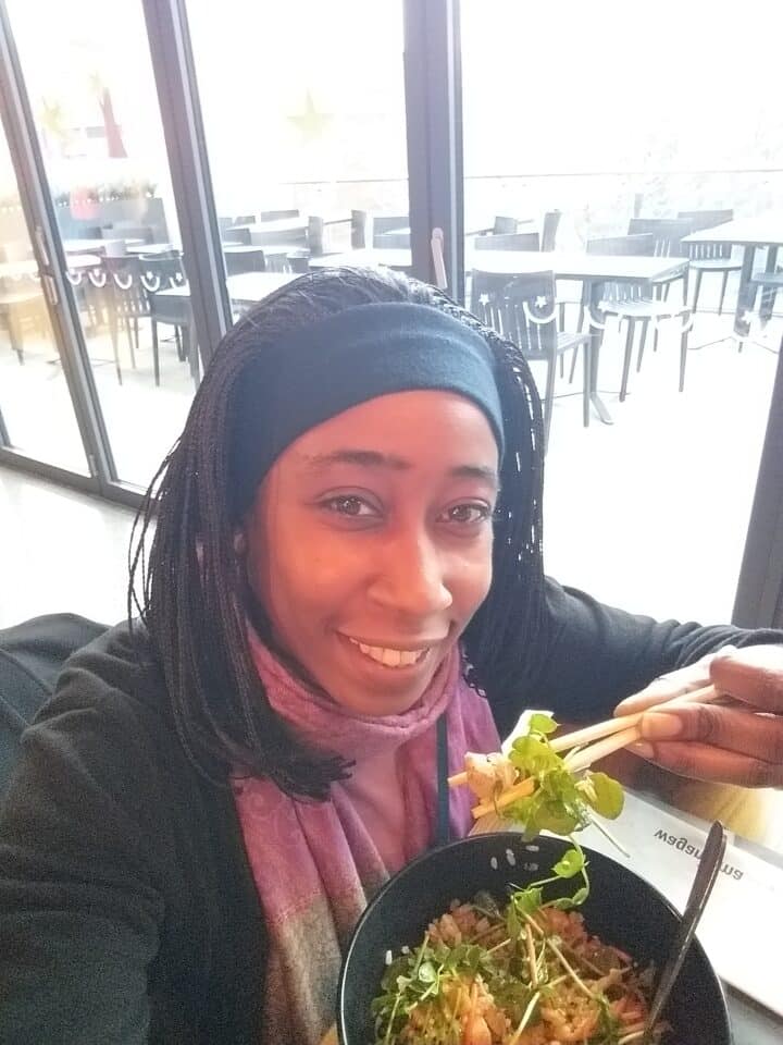 Smiling woman holding chopsticks with a bowl of rice and greens by a window.
