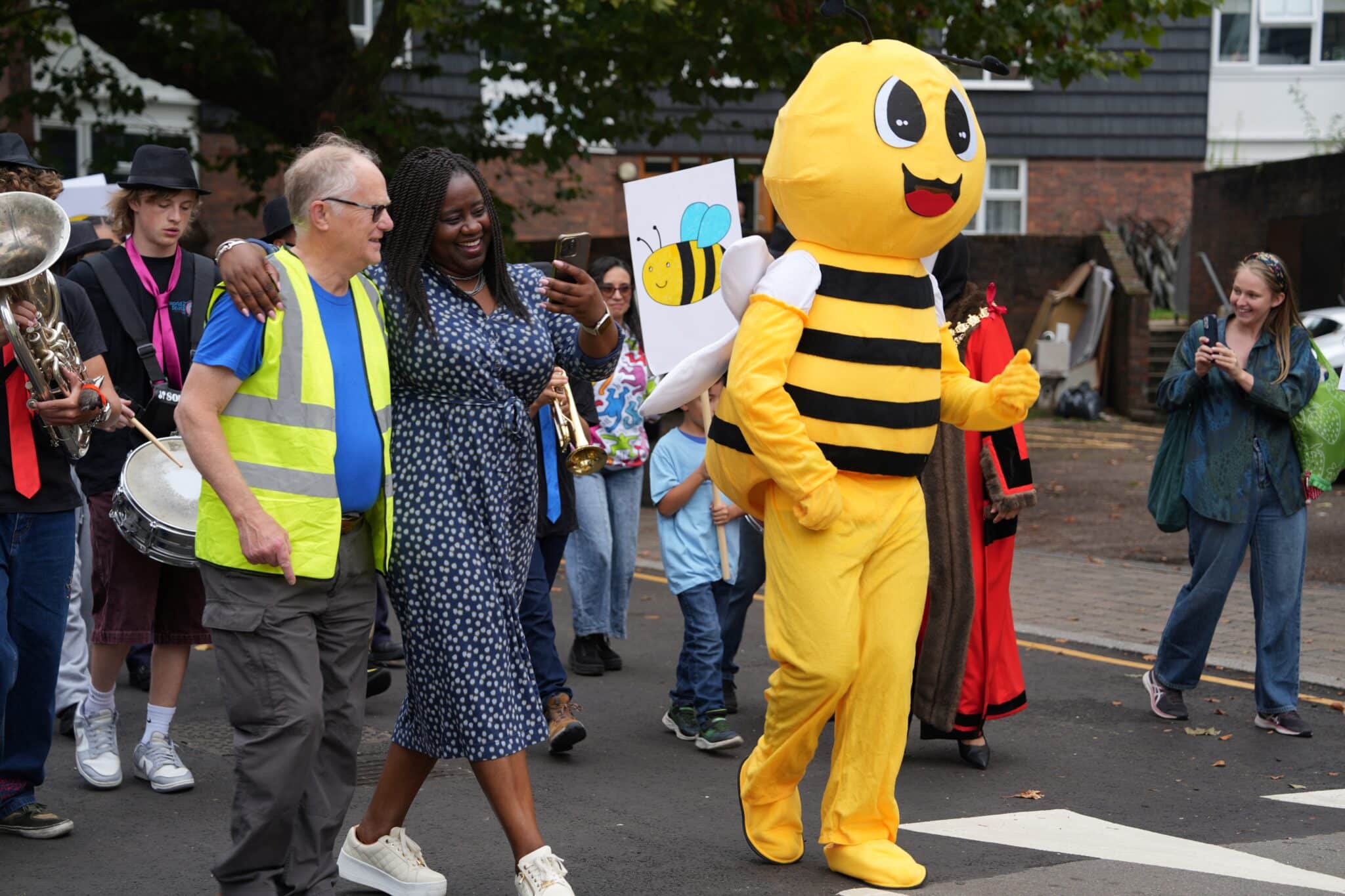 Community members participate in a joyful parade during the Battersea Community Festival 2024. A man in a yellow safety vest walks arm-in-arm with a woman taking a selfie, while a person in a cheerful bee costume leads the procession. Behind them, a band plays instruments and children hold a handmade bee sign. 