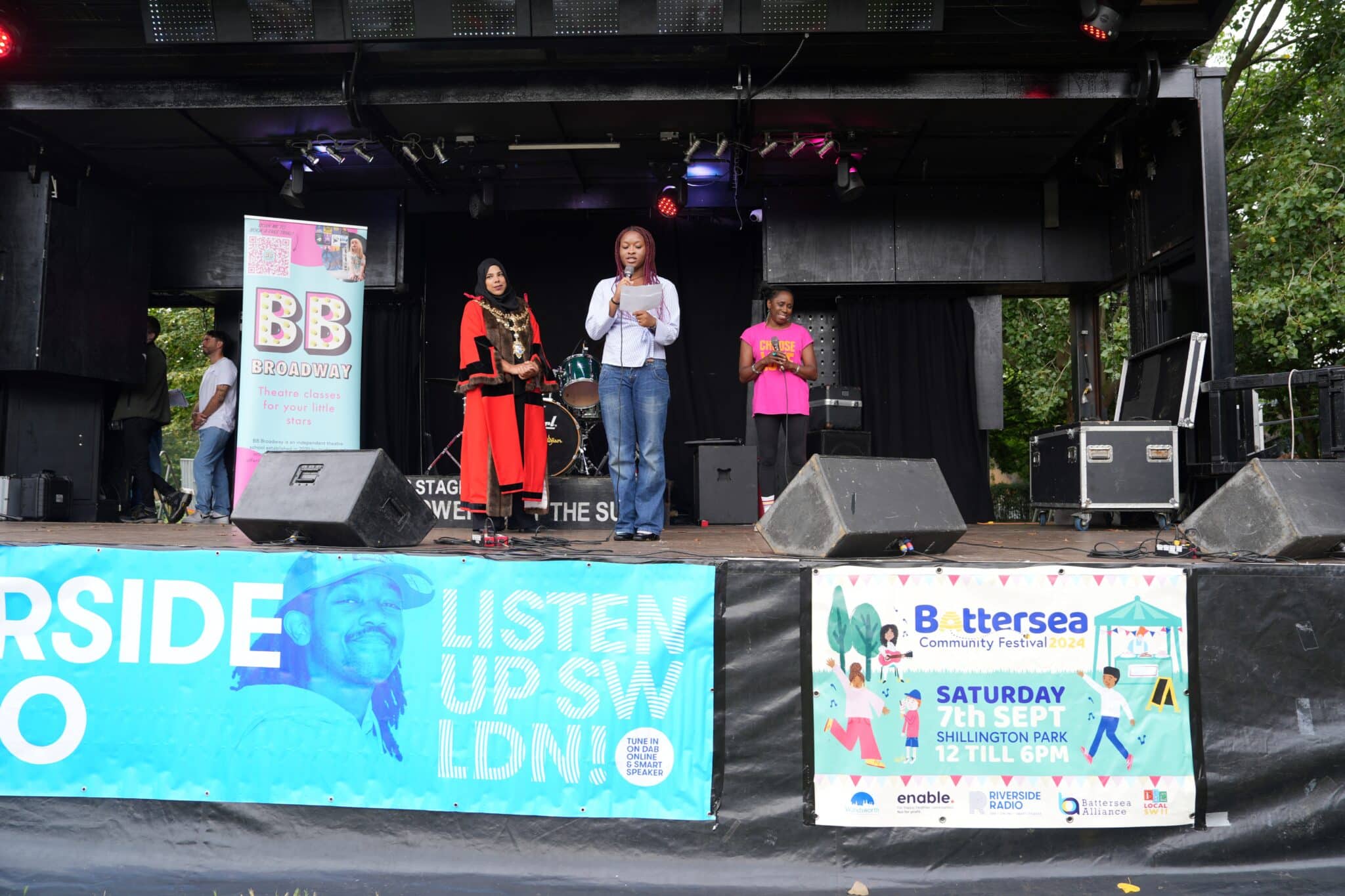 Three women stand on an outdoor stage at the Battersea Community Festival 2024 in Shillington Park, London. One is dressed in ceremonial mayoral robes, another reads from a paper, and a third wears a fitness-themed shirt. Behind them is a drum set and stage lighting.