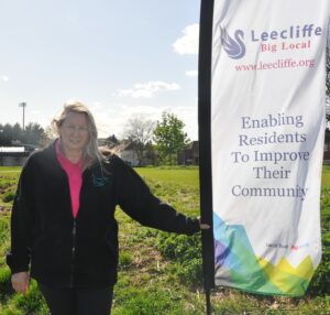 Donna Inwood stands outdoors on a grassy area next to a vertical banner. She is wearing a black fleece jacket over a pink polo shirt and has long blonde hair. The banner beside her reads: "Leecliffe Big Local www.leecliffe.org Enabling Residents To Improve Their Community" The banner also features a swan logo and colourful geometric designs at the bottom, along with the logos for Local Trust and Big Local. Trees, houses, and a sports field floodlight can be seen in the background under a bright, partly cloudy sky.