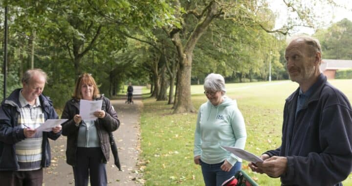 The image shows four adults standing outdoors on a tree-lined path in a park. They are holding papers and are engaged in a health walk. All are dressed in casual clothing suitable for mild weather. The mood appears lighthearted and relaxed, with some of the individuals smiling or laughing