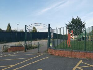 The image shows the entrance to "Meadow Park," as indicated by the arched metal sign above the gate. The entrance is flanked by green metal fencing, with a decorative orange silhouette of a person playing tennis attached to the fence on the right. There is a paved pathway leading into the park, and tennis courts are visible inside. 