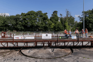 A group of people wearing orange high-visibility vests standing on or near a large railway turntable, used for turning locomotives. The turntable is surrounded by trees and safety barriers. Various signs and logos are displayed on the turntable, including Network Rail, DB Cargo, Historic England, MPower Kernow, and the Railway Heritage Trust.