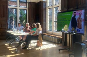 A panel of four people is seated at a table in a bright, wood-panelled room with large, leaded glass windows. Sunlight streams in, casting shadows on the wooden floor. To the right, a large screen displays a presentation slide titled "Community-led health initiatives: The practice, the potential and the challenges," part of the "Local Trust Research seminars."