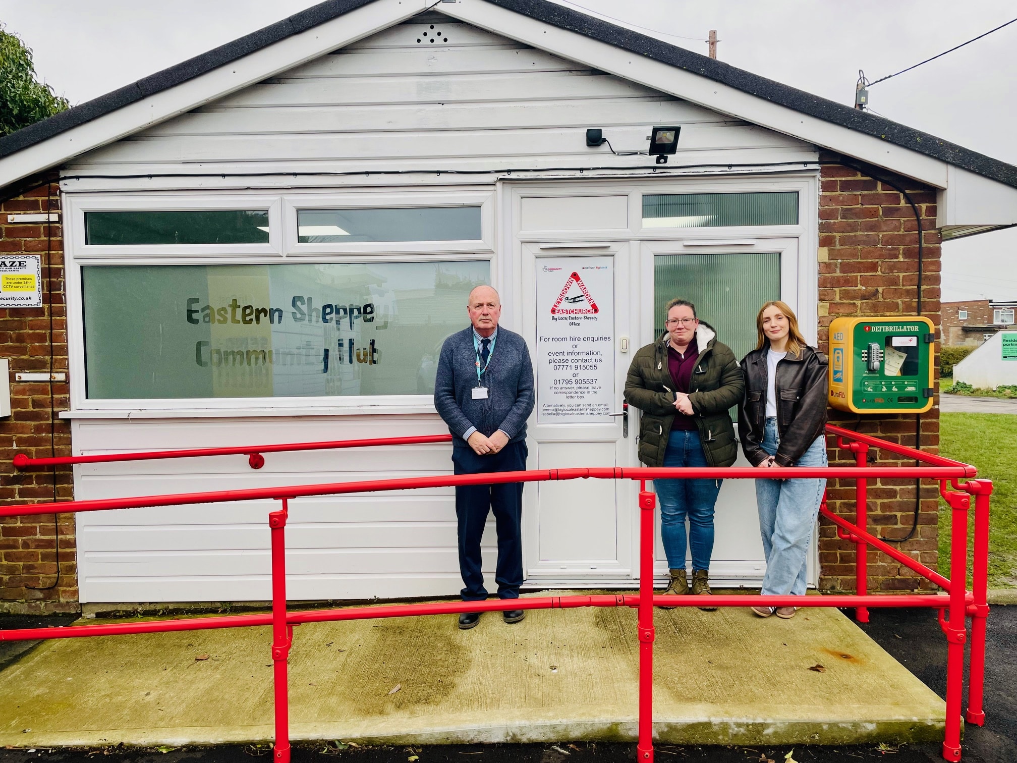 Three people standing outside the Eastern Sheppey Community Hub building. The building has a white front with a red handrail and accessibility ramp leading to the entrance. To the right of the door is a public defibrillator mounted on the wall.