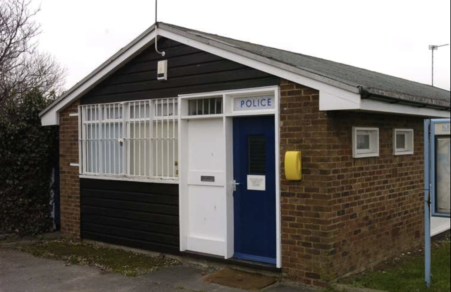 A small brick building with a sign above the door reading 'POLICE'. The front door is dark blue with a notice attached, and there are white bars on the windows next to it. A yellow box is mounted on the wall near the entrance.