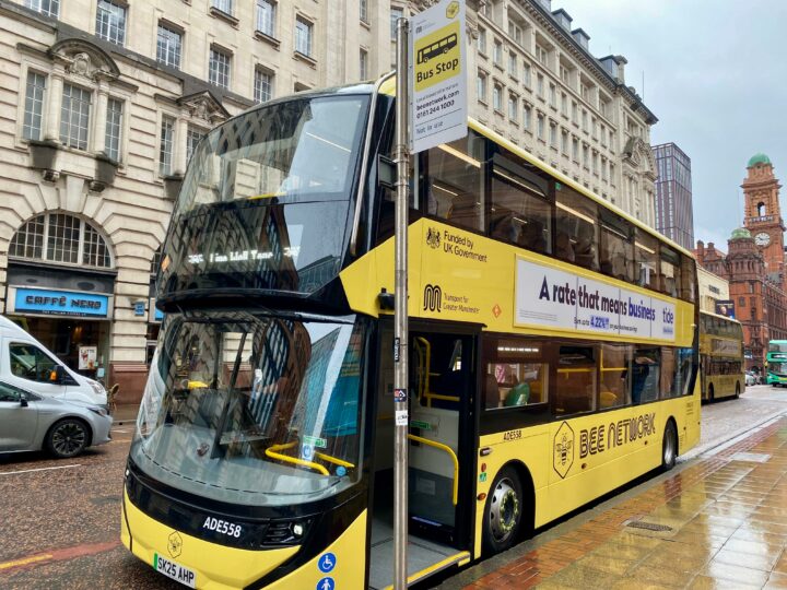 A bright yellow Bee Network double-decker bus parked on a wet city street in Manchester, branded with 'Funded by UK Government' and 'Transport for Greater Manchester'.