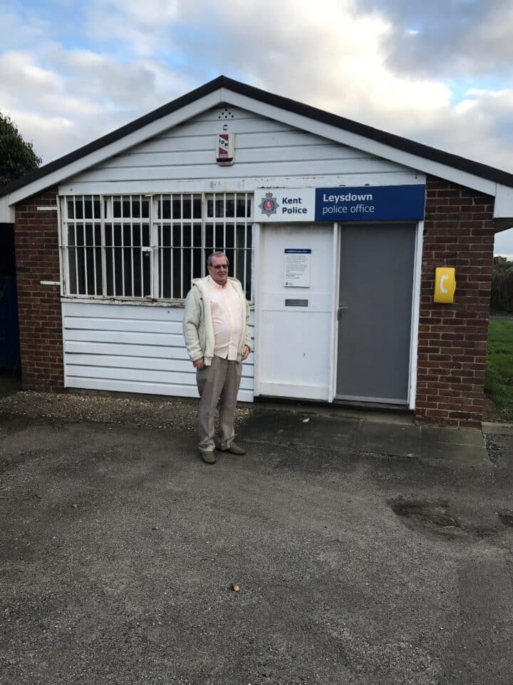 A man standing outside the Leysdown Police Office, part of Kent Police, on a cloudy day. The man is wearing glasses, a cream top and jacket, and grey trousers.