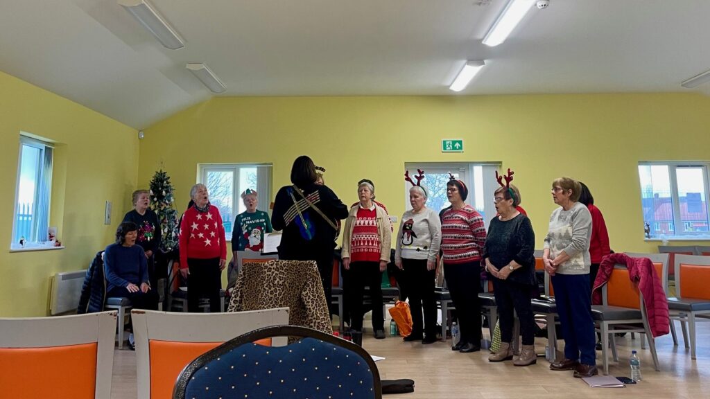 A choir group stand in a semi circle singing, one person stands in front with their back to the camera. The group of people are wearing Christmas jumpers and festive headwear.