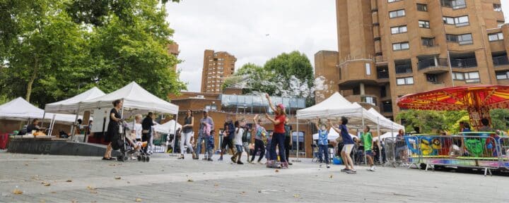 A vibrant community event scene. Children jump in the air as they try to catch huge bubbles, and their parents look on. There are white marquee tents, a colourful fairground ride and behind are some tall brick blocks of flats.