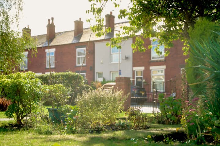 A terraced row of brick houses sits behind a lush green garden, with trees and hedges. It's a bright, sunny day.