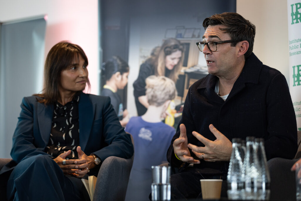 Two people sit and engage in discussion during a panel event. The woman on the left wears a blue suit and looks attentively toward the man speaking beside her, who is wearing glasses and a dark jacket. A banner with the logo “IPPR” (Institute for Public Policy Research) and a blurred background image of people working together can be seen behind them. Bottles of water and a paper cup sit on the table in front.