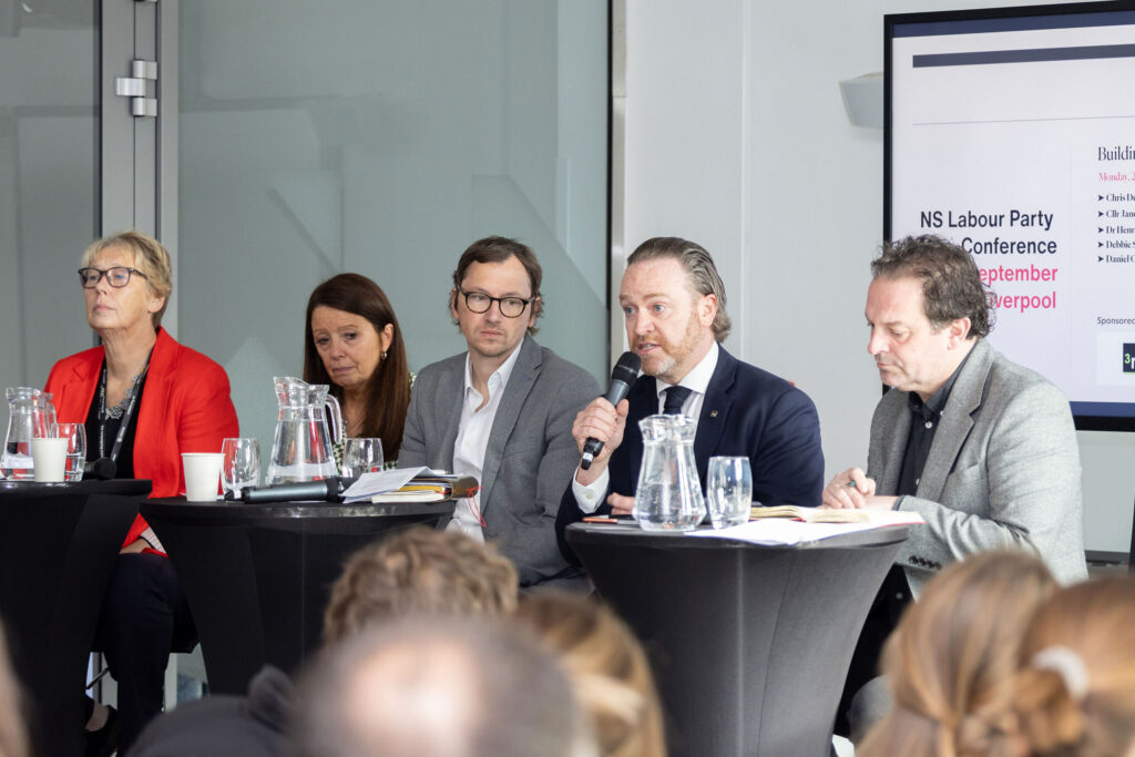 A panel of five people sits at tables during a conference discussion. One man holds a microphone and speaks while others listen attentively. Papers, glasses, and water pitchers are on the tables. A presentation screen in the background displays text. The audience is visible in the foreground.