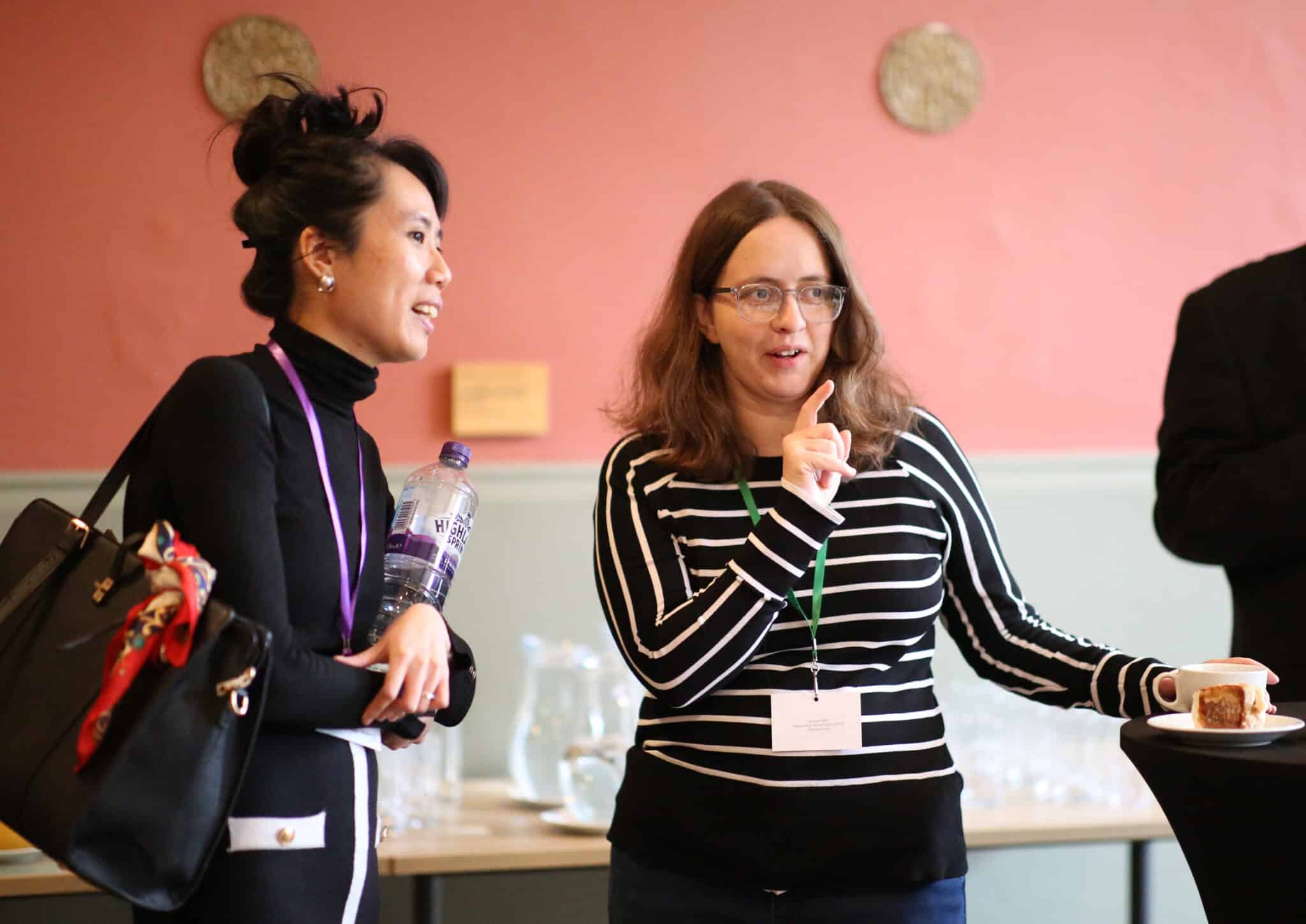 Two attendees chatting at the All Together Now conference. They both wear event lanyards. Behind them is a bright pink wall.