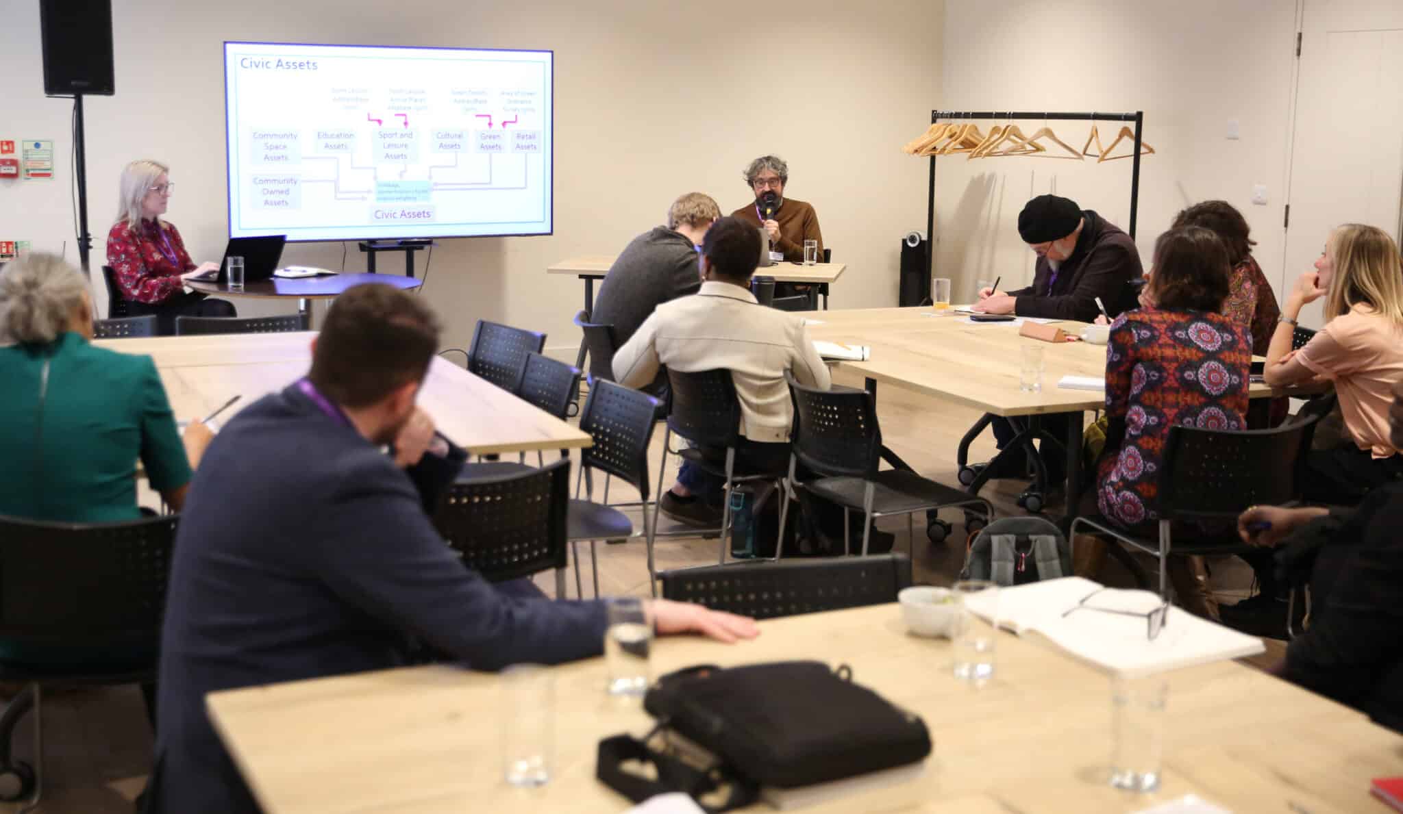 Groups of people sit at rectangular tables for a breakout session at a conference. They face the front of the room where two people lead the sessions. A whiteboard reads 'Civic assets' with a flow chart diagram.