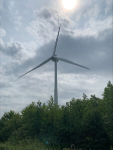 A tall white wind turbine rises above a line of green trees, its three blades extending against a cloudy sky. The sun is visible through thin clouds near the top of the frame, casting soft light over the scene.