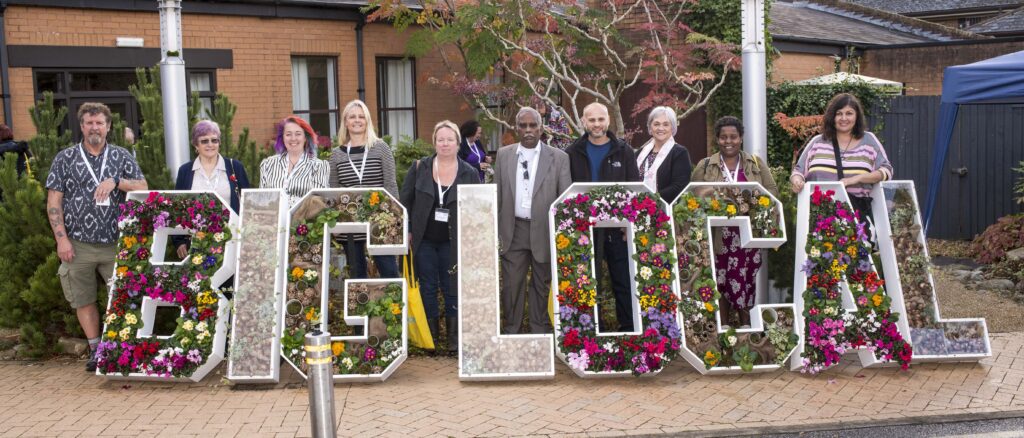 A group of adults stand outdoors behind large freestanding letters spelling “BIG LOCAL,” each letter filled with colourful flowers and plants. The group faces the camera in a paved courtyard with buildings and greenery in the background.