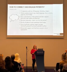 A woman in a red top stands at a lectern speaking to an audience at a conference, with a slide projected behind her titled “HOW TO CONNECT AND ENGAGE PATIENTS?” 