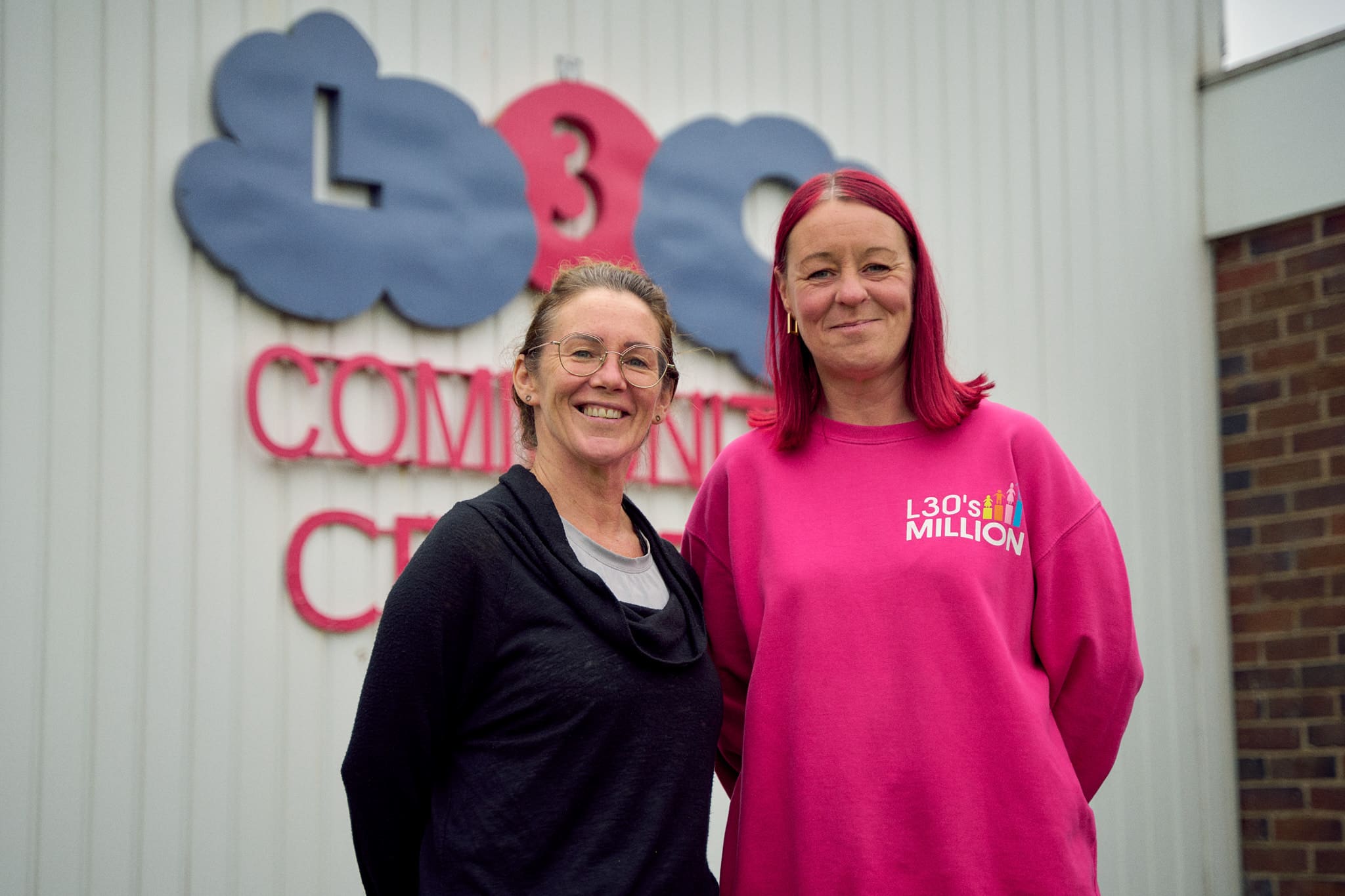 Two women stand in front of a community centre with a pink and purple sign that reads 'L30 community centre'.