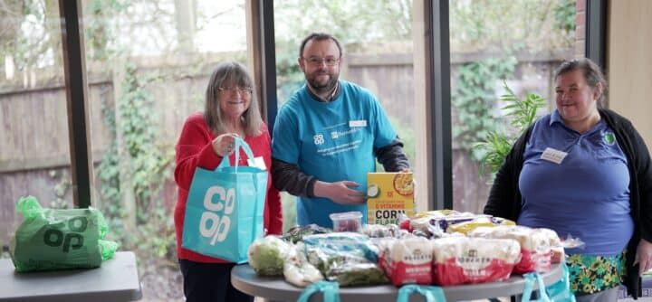 Three adults stand behind a round table covered with groceries including bread, vegetables and a box labelled “Corn Flakes,” while one woman holds a turquoise shopping bag with the white “Co op” logo; the man in the centre wears a blue T-shirt reading “Co op” and “Barnardo’s – Bringing communities together to support young people.