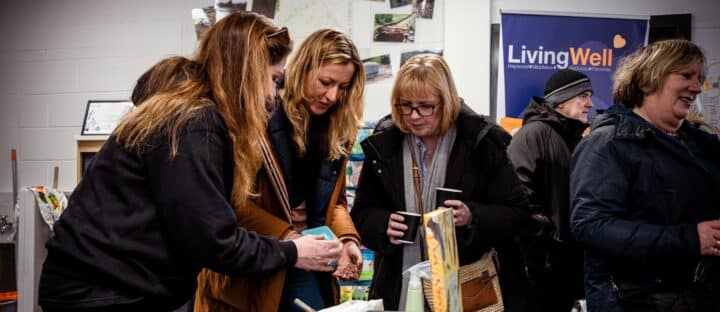 Three women look at items on a table at a community event, with a LivingWell banner in the background