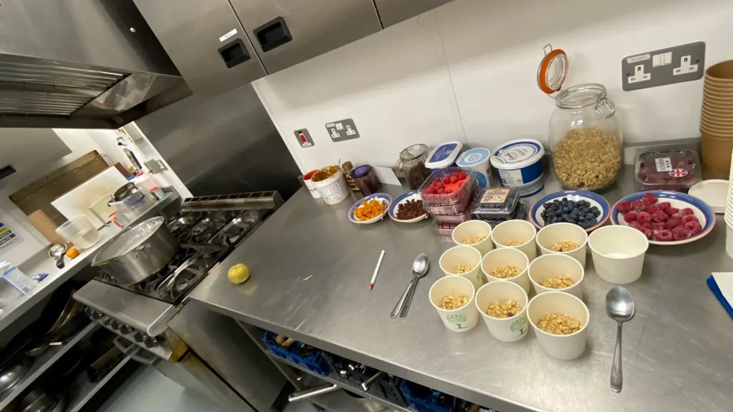 A kitchen preparation area with bowls of granola arranged on a stainless-steel counter, alongside fresh berries, yoghurt tubs and jars of oats. A large pot sits on the stove nearby.
